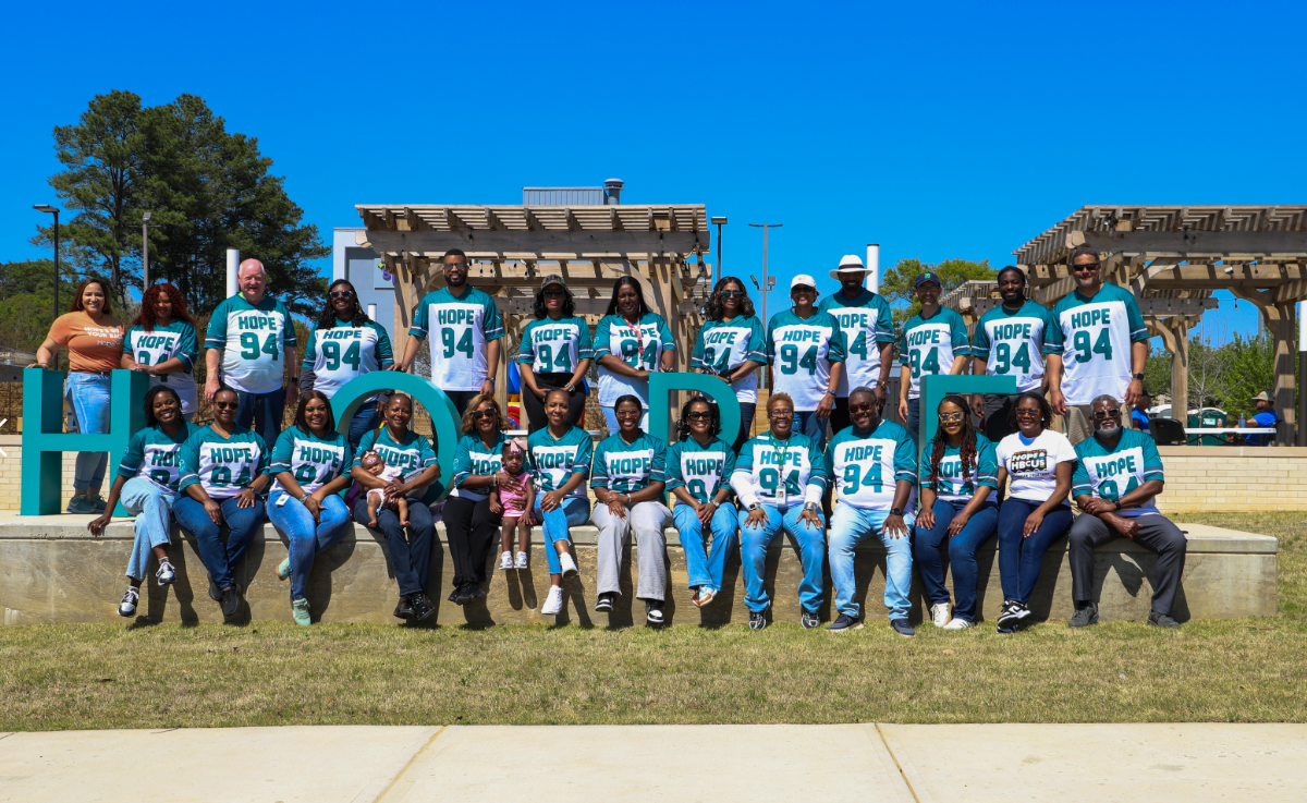 A large group of HOPE team members and community participants pose outdoors in a park on a sunny day, many wearing teal “HOPE 94” shirts. They are arranged in two rows around large decorative letters spelling “HOPE,” with wooden pergolas and trees in the background.