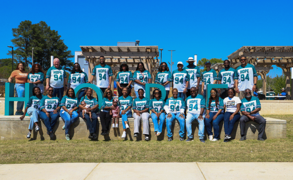 A large group of HOPE team members and community participants pose outdoors in a park on a sunny day, many wearing teal “HOPE 94” shirts. They are arranged in two rows around large decorative letters spelling “HOPE,” with wooden pergolas and trees in the background.