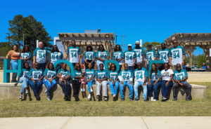 A large group of HOPE team members and community participants pose outdoors in a park on a sunny day, many wearing teal “HOPE 94” shirts. They are arranged in two rows around large decorative letters spelling “HOPE,” with wooden pergolas and trees in the background.